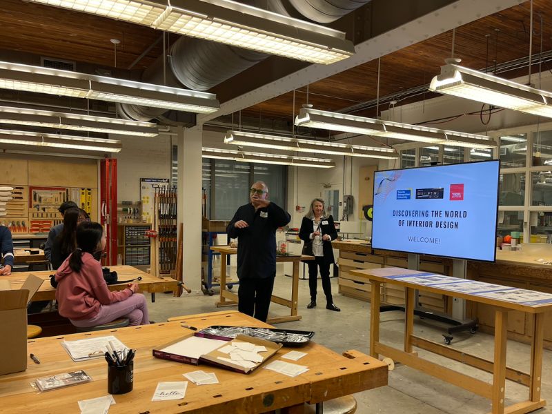 A man standing beside a TV and instructing in a workshop looking classroom in front of multiple students. 